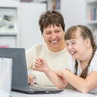 Smiling girl with down syndrome is uses a laptop with her teacher at library. Education for disabled children concept.